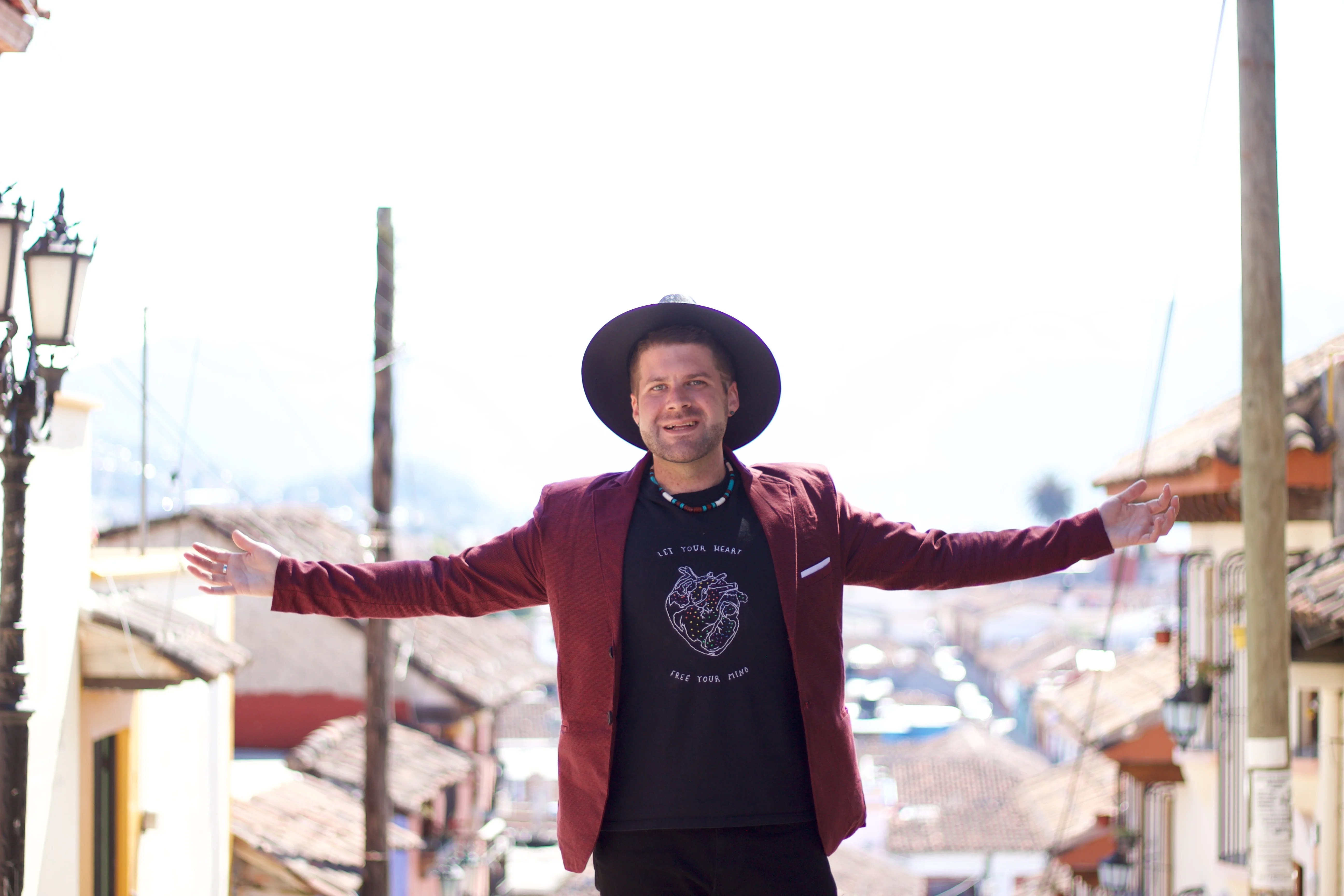 Grant James is standing on top of a staircase in Mexico with his arms stretched out. Wearing a black shirt, burgundy blazer and wide brimmed black hat.