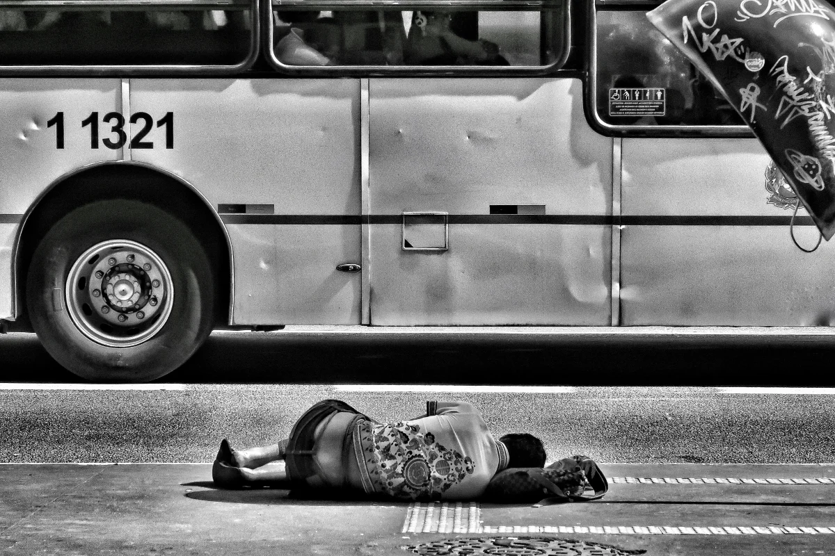 Homeless person sleeping on street in front of bus, black and white image.