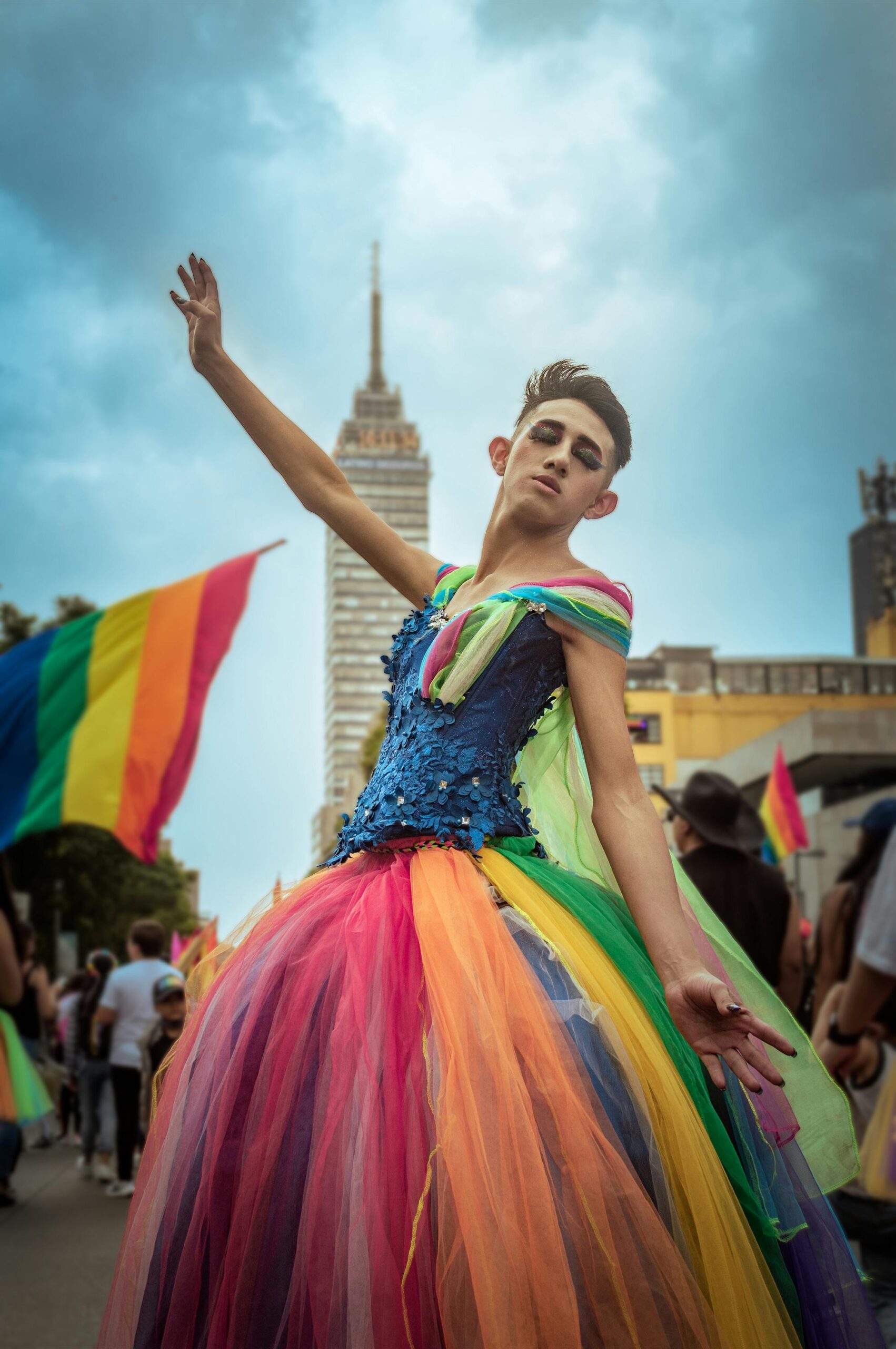 LGBTQ+ person in rainbow skirt in front of a tower looking happy and free.